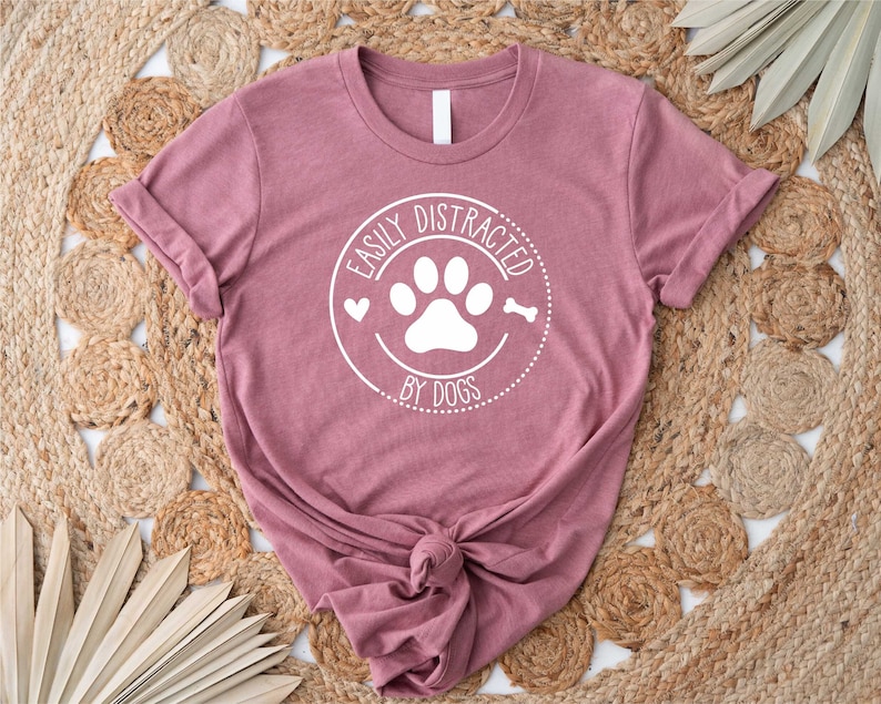 May include: A mauve t-shirt with a white circle graphic that says "Easily Distracted by Dogs" with a paw print and bone inside the circle.