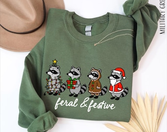 May include: A military green crewneck sweatshirt featuring a graphic of four raccoons. One is wrapped in Christmas lights, one holds a gift, one wears a gingerbread costume, and one wears a Santa suit. The text reads feral & festive.