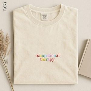 May include: An ivory short-sleeved t-shirt with a crew neckline. The shirt features the embroidered text "occupational therapy" in multicolored lowercase letters centered on the chest. The interior neck label reads "Comfort Colors".