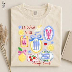 a beige t-shirt with a colorful graphic design depicting various food items and icons, including fish, a lobster, a lemon, and a bowl of fruit.