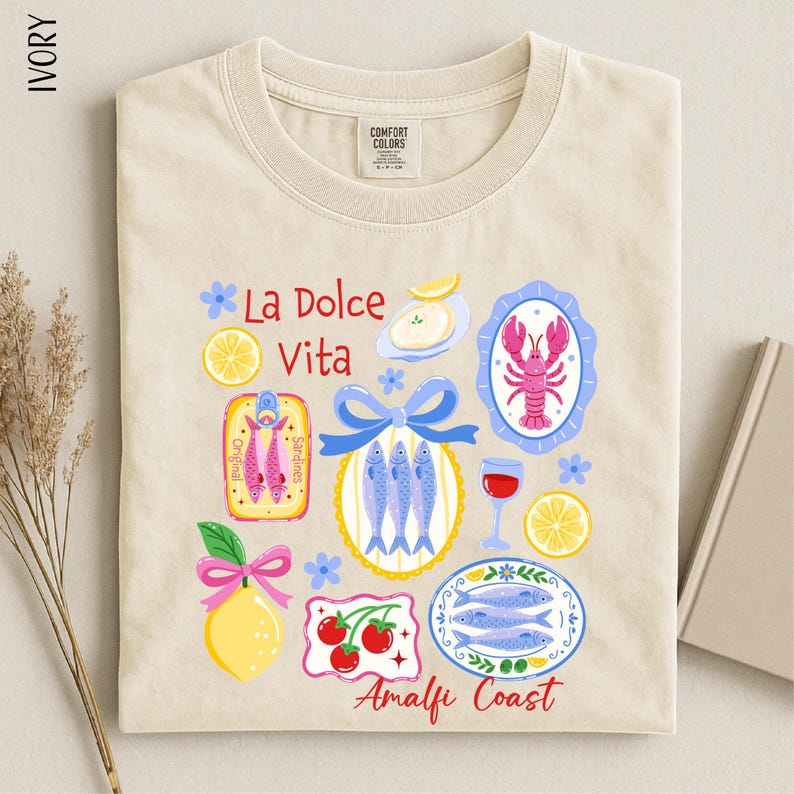 a beige t-shirt with a colorful graphic design depicting various food items and icons, including fish, a lobster, a lemon, and a bowl of fruit.
