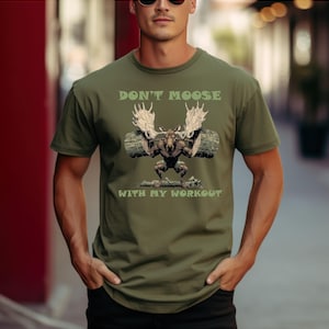 Könnte beinhalten: Olivgrünes T-Shirt mit dem Text "DON'T MOOSE WITH MY WORKOUT". Die Grafik zeigt einen Elch beim Gewichtheben. Der Elch hat große Geweihe und befindet sich in einer Hockposition. Text und Grafik sind in Grün- und Brauntönen gehalten.