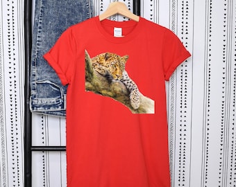 Puede incluir: Una camiseta roja con una ilustración detallada de un leopardo descansando sobre una rama de árbol. El leopardo tiene un pelaje manchado y está acostado. La camiseta está colgada en una percha de madera.