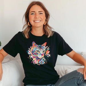 Może przedstawiać: Czarny t-shirt z krótkim rękawem z żywym, wielokolorowym nadrukiem graficznym przedstawiającym pysk kota na klatce piersiowej. Ilustracja kota wykorzystuje abstrakcyjne kształty i jasne odcienie, w tym różowy, niebieski, żółty i pomarańczowy.