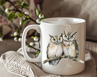 May include: White ceramic mug featuring a watercolor illustration of two owls perched on a branch. The owls have brown, white, and yellow feathers. The mug has a large handle and is sitting on a beige knitted blanket.