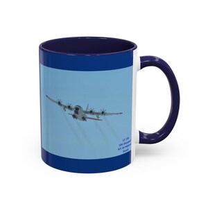 Könnte beinhalten: Eine weiße Keramiktasse mit blauem Innenraum und Henkel. Die Tasse zeigt ein Foto eines weißen Flugzeugs, das vor einem hellblauen Himmel fliegt. Der Text "LC-130 (Ski-Equipped) NY Air National Guard" ist auf die Tasse gedruckt.