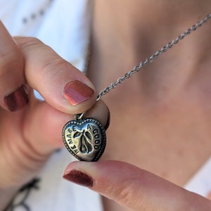 May include: A silver heart-shaped pendant necklace with the words "We make a good" engraved on it. The pendant features a small pear design.