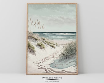 May include: A framed print of a beach scene with a path leading through the sand dunes to the ocean. The sky is a soft blue and the water is a turquoise color. The sand is white and there are footprints in the sand. The print is framed in a light wood frame.