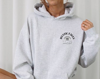 Pode incluir: Uma sudadera com capuz cinza com uma estampa gráfica preta de uma boca de vampiro com presas e o texto "Mystic Falls Social Club" na frente.