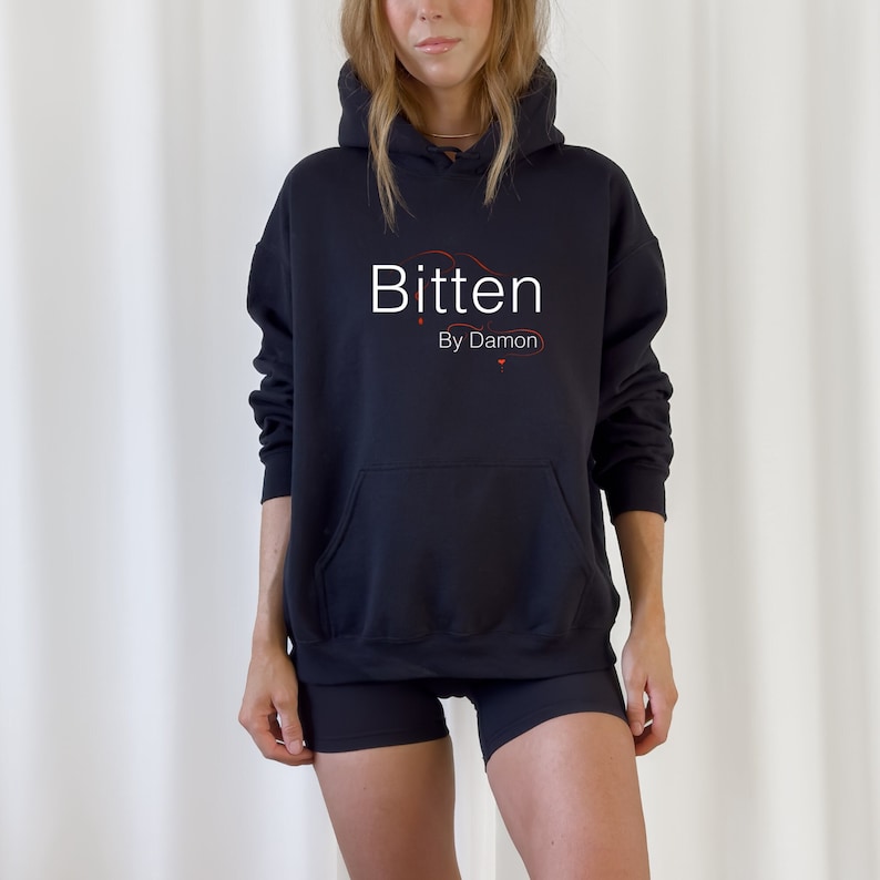 May include: Black hoodie with the text "Bitten by Damon" in white and red. The hoodie has a front pocket and a drawstring hood. The model is wearing black shorts.