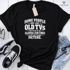 May include: A black short-sleeved t-shirt featuring white text that reads: SOME PEOPLE ARE LIKE OLD TVs THEY NEED TO BE SLAPPED A FEW TIMES TO GET THE F*CKING PICTURE. The shirt has a crew neckline and a white tag attached to the collar area.