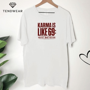 Puede incluir: Una camiseta blanca de manga corta con el texto en rojo oscuro Karma is like 69: You get, what you give impreso en el centro del pecho. La camiseta tiene un cuello redondo y se muestra en una percha de madera sobre un fondo blanco.