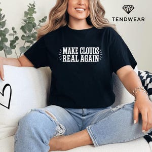 Op de afbeelding: Zwart T-shirt met de witte tekst "MAKE CLOUDS REAL AGAIN". Het shirt heeft een ronde hals en wordt door een persoon gedragen. Het merklogo "TENDWEAR" is rechtsboven zichtbaar.