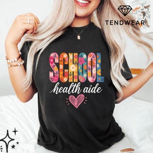 Puede incluir: Camiseta negra con las palabras "SCHOOL health aide" en un diseño floral. Un corazón rosa con brillos está debajo del texto. La camiseta es de cuello redondo.