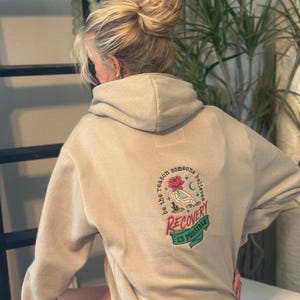 Può includere: Una felpa con cappuccio color beige caratterizzata da un ricamo sul retro. Il disegno raffigura una mano che tiene un fiore rosa, circondata dal testo "be the reason someone believes" e "RECOVERY IS POSSIBLE" su un nastro verde.