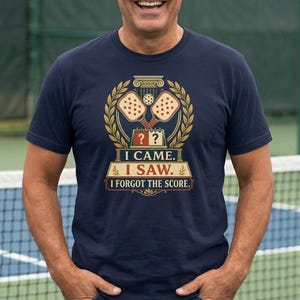 May include: A navy blue short-sleeved t-shirt featuring a graphic print of two pickleball paddles, a ball, and a scoreboard. The text reads I CAME. I SAW. I FORGOT THE SCORE. The design is framed by a golden laurel wreath and architectural columns.