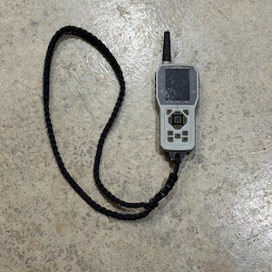 May include: A gray and black handheld device with a small antenna and a black strap. The device has a screen with a cracked display and a keypad with several buttons.