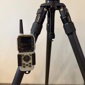 May include: A black tripod with a remote control attached to one of the legs. The remote control has a small screen and several buttons. The tripod is made of carbon fiber and has a black finish.