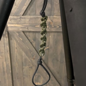 Puede incluir: Un cordón de paracord negro y verde oliva con un lazo. El cordón está sujeto a un anillo de metal. La sección verde oliva está trenzada. El fondo es una puerta de madera.