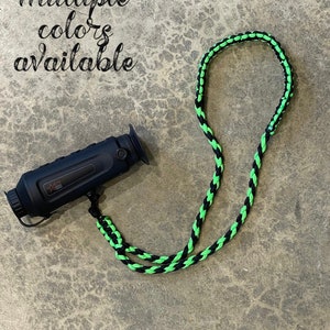 Könnte beinhalten: Ein geflochtenes Seilband in Schwarz und Grün mit einem schwarzen Monokular daran. Das Band ist aus Paracord und soll als Halsband oder Schulterband verwendet werden. Das Monokular ist ein kleines, handliches Teleskop, das zum Beobachten von Vögeln, zur Jagd oder für andere Outdoor-Aktivitäten verwendet werden kann.  "Mehrere Farben verfügbar" steht in schwarzer Schrift auf grauem Hintergrund.