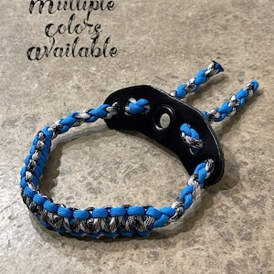 Puede incluir: Una correa de muñeca trenzada azul y gris con un lazo de cuero negro y un anillo de metal. Múltiples colores disponibles.