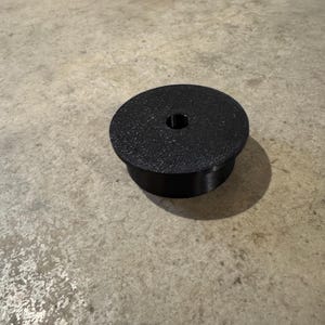 May include: A black, circular object with a textured surface and a central hole. The object has a cylindrical base and sits on a concrete surface. The object appears to be made of plastic or a similar material.