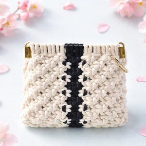 Puede incluir: Una bolsa cuadrada de macramé blanquecino con una franja negra central. La bolsa tiene un diseño tejido texturizado y herrajes metálicos dorados. Flores y pétalos rosas están esparcidos alrededor de la bolsa.