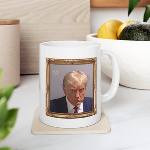 May include: White ceramic mug with a gold-framed image of a man with blonde hair. The man is wearing a dark suit and a red tie. The image is on a light gray background.