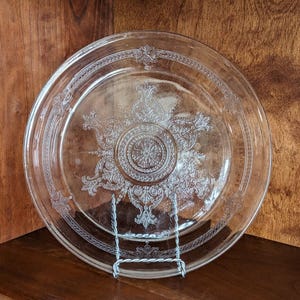May include: Clear glass plate with ornate, embossed floral and scrollwork design. The plate is round and sits on a silver metal stand. The plate is displayed on a dark wood surface.