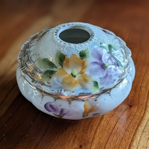 May include: A white porcelain trinket box with a floral design. The box has a round shape with a raised rim and a small opening in the center of the lid. The flowers are painted in shades of yellow, purple, and green.