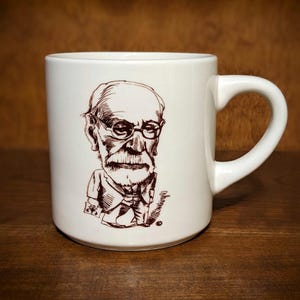 Puede incluir: Taza de cer&aacute;mica blanca con una ilustraci&oacute;n en tinta marr&oacute;n de un hombre con gafas y barba. La taza tiene un asa curvada y un acabado brillante. La ilustraci&oacute;n es una caricatura. La taza est&aacute; sobre una superficie de madera.