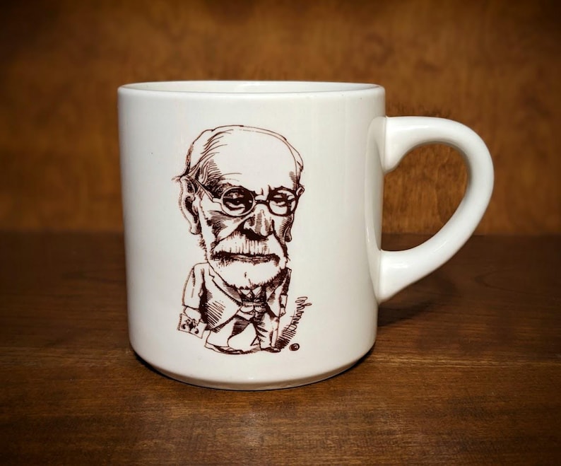 Puede incluir: Taza de cer&aacute;mica blanca con una ilustraci&oacute;n en tinta marr&oacute;n de un hombre con gafas y barba. La taza tiene un asa curvada y un acabado brillante. La ilustraci&oacute;n es una caricatura. La taza est&aacute; sobre una superficie de madera.