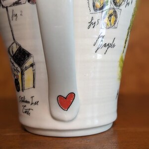 May include: White ceramic mug with a handle featuring hand-drawn illustrations. The mug has a red heart on the handle and drawings of Italian ice carts and bagels. The word "Bagels" is written in cursive.