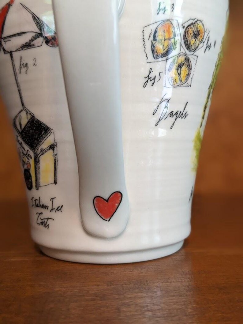 May include: White ceramic mug with a handle featuring hand-drawn illustrations. The mug has a red heart on the handle and drawings of Italian ice carts and bagels. The word "Bagels" is written in cursive.