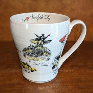 May include: White ceramic mug featuring illustrations of New York City landmarks, including Grand Central Terminal and a yellow taxi. The mug has a curved handle and the words "I love New York City" written in black script.