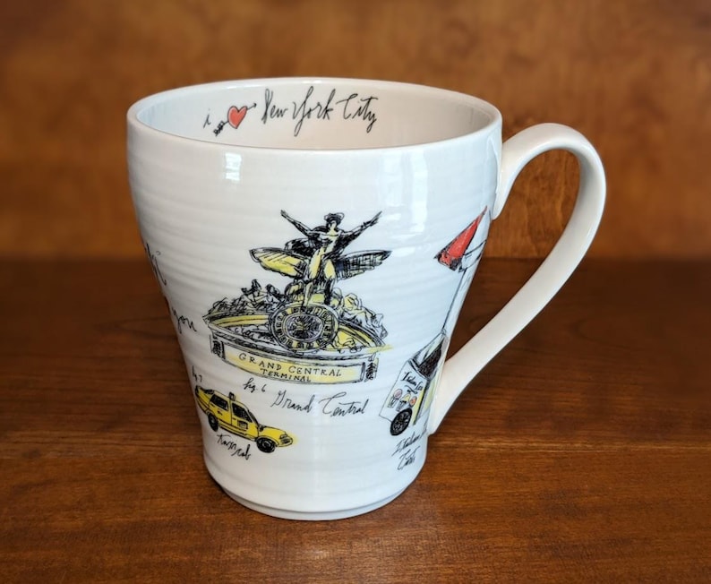 May include: White ceramic mug featuring illustrations of New York City landmarks, including Grand Central Terminal and a yellow taxi. The mug has a curved handle and the words "I love New York City" written in black script.
