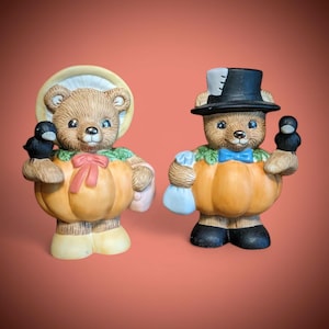 Könnte beinhalten: Zwei Keramik-Bärenfiguren. Einer der Bären trägt eine Haube und hält einen schwarzen Vogel, der andere trägt einen Zylinder und hält einen schwarzen Vogel. Beide Bären sind in Kürbiskostümen mit grünen und orangefarbenen Akzenten gekleidet.