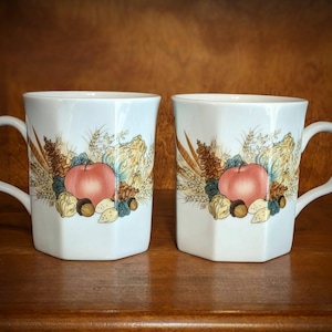 May include: Two white ceramic mugs with a handle, featuring a fall harvest design. Each mug has a red apple, nuts, and wheat stalks. The mugs are set on a wooden surface.