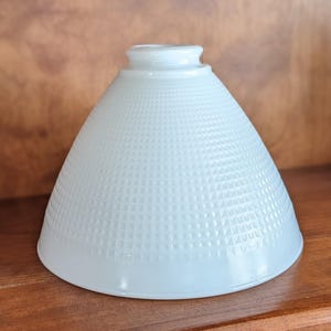 May include: A white glass lamp shade with a conical shape and a textured grid pattern of small raised squares across the exterior surface. The shade features a smooth, circular opening at the top for mounting and a wide, flared base opening.