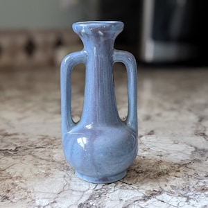 Pode incluir: Um vaso de cerâmica azul pequeno com duas alças. O vaso tem um esmalte azul claro manchado.
