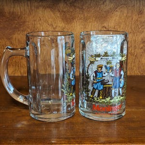 May include: Two clear glass beer mugs with handles. One mug is empty, the other has a colorful graphic of people in traditional clothing, the text "Metzger-Wirth" and "Moringo" in red. The mugs are on a wooden surface.