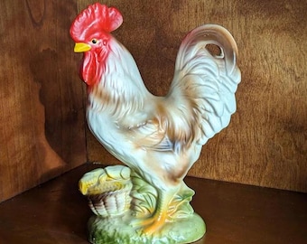 Pode incluir: Estatueta de galo de cerâmica com crista e barbela vermelhas, bico amarelo e penas brancas e castanhas. O galo está sobre uma base verde com uma cesta. A figura é um item decorativo.