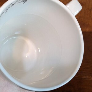 May include: A white ceramic mug with a square handle. The interior of the mug is smooth and glossy. The words "New York City" are written in a gray script on the side of the mug.