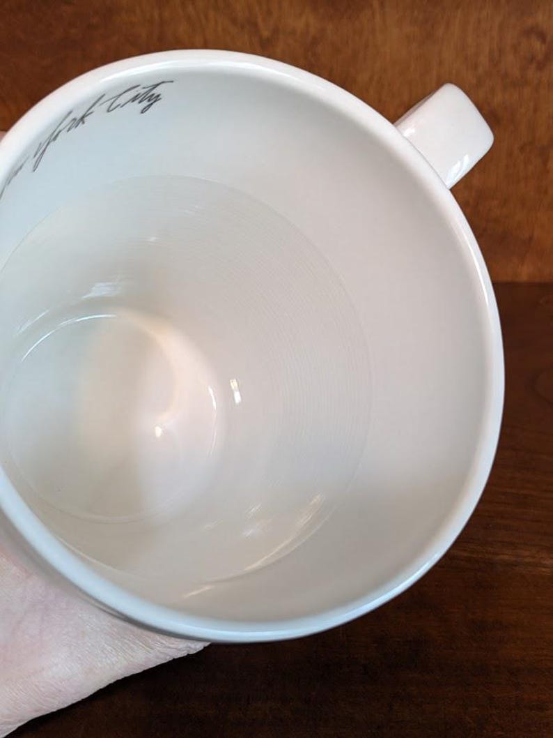 May include: A white ceramic mug with a square handle. The interior of the mug is smooth and glossy. The words "New York City" are written in a gray script on the side of the mug.