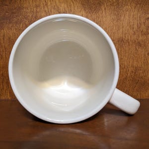Puede incluir: Una taza de cer&aacute;mica blanca con asa, vista desde arriba. El interior de la taza es de color crema claro, y el asa est&aacute; en el lado derecho. La taza est&aacute; sobre una superficie de madera marr&oacute;n oscuro.