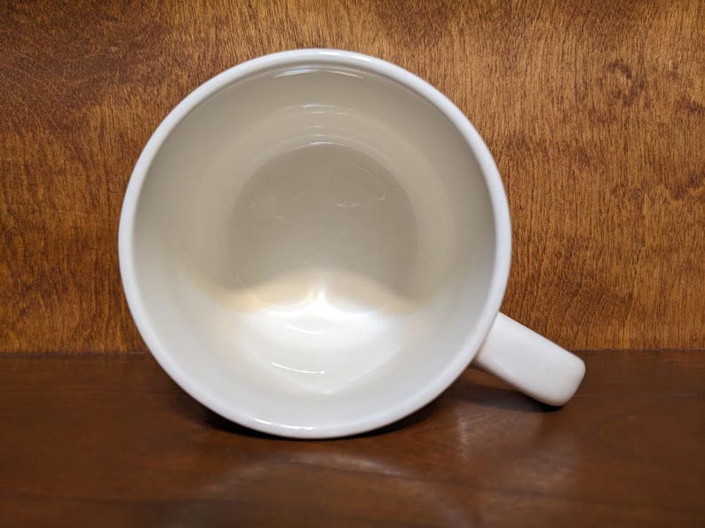 Puede incluir: Una taza de cer&aacute;mica blanca con asa, vista desde arriba. El interior de la taza es de color crema claro, y el asa est&aacute; en el lado derecho. La taza est&aacute; sobre una superficie de madera marr&oacute;n oscuro.