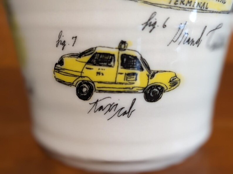 May include: White ceramic mug with a yellow taxi cab illustration. The taxi has black wheels and windows, with the word "taxicab" written below. The mug also has handwritten text, including "fig. 6" and "fig. 7".