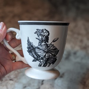 May include: White ceramic mug with a black floral design. The mug has a footed base.