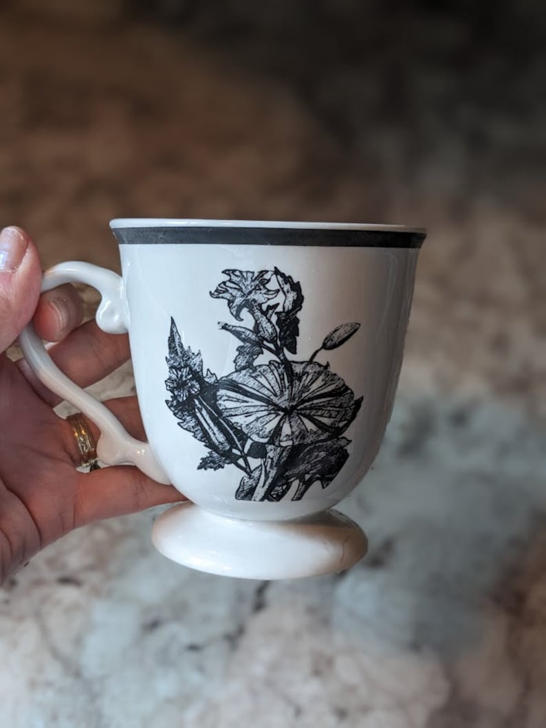 May include: White ceramic mug with a black floral design. The mug has a footed base.