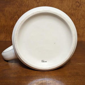 Puede incluir: Una taza de cer&aacute;mica blanca, vista desde abajo, con una base redondeada y un asa peque&ntilde;a. La palabra "China" est&aacute; impresa en escritura negra cerca del centro. La taza est&aacute; sobre una superficie de madera oscura.